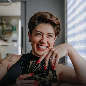 May include: A person with short brown hair is smiling and holding a deck of black tarot cards with gold accents. The cards are fanned out and show the backs of the cards. The person is wearing a black tank top.