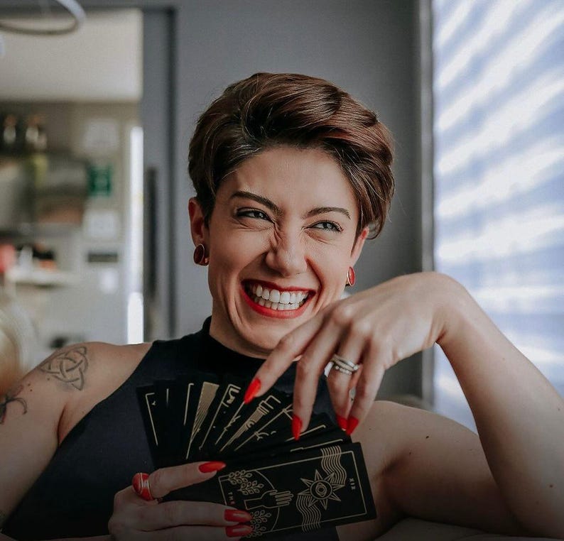 May include: A person with short brown hair is smiling and holding a deck of black tarot cards with gold accents. The cards are fanned out and show the backs of the cards. The person is wearing a black tank top.
