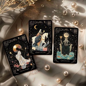 Pode incluir: Tr&ecirc;s cartas de tar&ocirc; com fundos pretos e detalhes dourados. As cartas apresentam ilustra&ccedil;&otilde;es de figuras e elementos celestes. Uma carta diz "Two of Wands", outra "Knight of Cups" e a terceira "King of Cups".