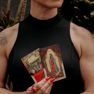 May include: Two tarot cards, one with a skeleton figure wearing a hat and the other with a woman in a red dress and a halo. The cards are held in the hands of a person wearing a black tank top.