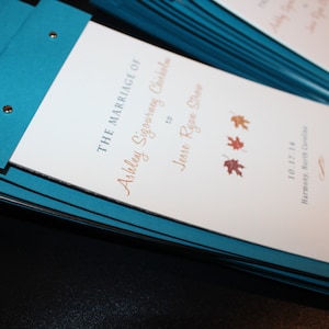 Könnte beinhalten: Ein Stapel blauer Programmhefte für eine Hochzeitszeremonie. Der Einband zeigt den Text "THE MARRIAGE OF" gefolgt von den Namen "Ashley Sigourney Chisholm" und "Jesse Ryan Shoe" in einer Schreibschrift. Das Datum "10.17.14" und der Ort "Harmony, North Carolina" sind unter den Namen gedruckt.