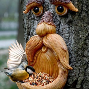 Puede incluir: Un comedero para pájaros caprichoso con forma de rostro tallado en un tronco de árbol. El rostro tiene una larga barba y está lleno de alpiste. Un pequeño pájaro con plumas amarillas, negras y blancas come del comedero. El rostro es de color marrón claro.