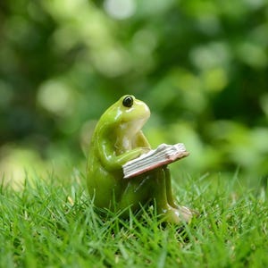 以下が含まれることがあります： 緑色のカエルの置物が、赤い縁取りのページがある本を読んで、緑の草の上に座っています。カエルは黒い目を持ち、ぼやけた緑色の背景を背景に配置されており、庭の環境を示唆しています。