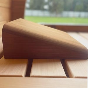 Può includere: Un poggiatesta per sauna in legno con la parte superiore curva e un design angolato. Il poggiatesta è realizzato in legno marrone chiaro e poggia su una panca in legno a doghe. Lo sfondo è sfocato, mostrando una vista sul verde.