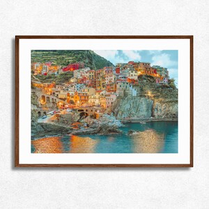 以下が含まれることがあります： 夕暮れ時のイタリアの海岸沿いの村の額入りプリント。カラフルな建物が丘を下って海に向かう様子が描かれています。水面には村の明かりが映り、温かい雰囲気を作り出しています。フレームは濃い茶色です。