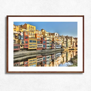 Puede incluir: Una impresión enmarcada de un paisaje urbano europeo. Edificios coloridos bordean un río, reflejándose en el agua. Los edificios están pintados en tonos de amarillo, naranja, rosa y azul. El cielo es claro y azul.