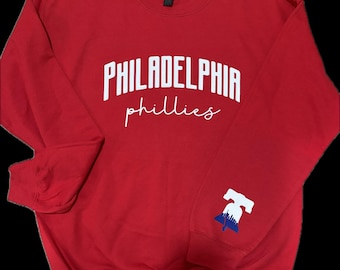 May include: A red crewneck sweatshirt with the words "PHILADELPHIA phillies" in white block and script lettering. A Liberty Bell graphic with blue and white accents is on the sleeve. The sweatshirt is a solid red color.