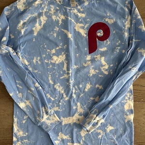 May include: A light blue long-sleeved t-shirt with a white tie-dye pattern and a red "P" on the front.