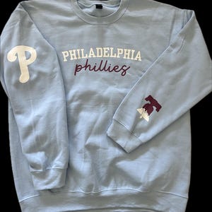 Könnte beinhalten: Hellblauer Pullover mit den Worten "PHILADELPHIA phillies" in Weiß und Maroon auf der Vorderseite. Der linke Ärmel hat ein weißes "P" und der rechte Ärmel eine Liberty Bell in Maroon.