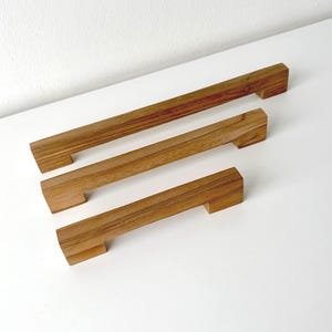 Peut inclure: Trois poignées de placard rectangulaires en bois disposées en diagonale, de la plus longue à la plus courte. Chaque poignée présente un motif de grain de bois brun naturel avec une finition lisse et deux blocs de montage carrés aux extrémités.