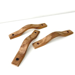 Può includere: Tre maniglie per armadietti in legno curvato con una venatura marrone naturale. Ogni maniglia presenta un design liscio e arcuato con due fori di montaggio circolari preforati alle estremità, che mostrano la consistenza del legno.