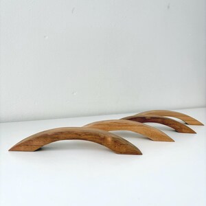 May include: Three arched cabinet pulls crafted from natural wood with a visible grain pattern. Each handle features a smooth, curved design with tapered ends, showcasing a warm brown tone and a polished finish for furniture hardware applications.