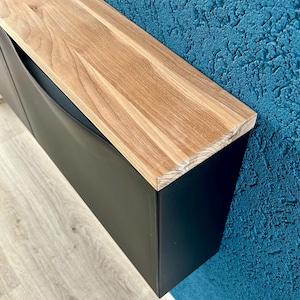 Könnte beinhalten: Ein moderner schwarzer Schrank mit einer hellbraunen Holzplatte. Der Schrank hat ein schlankes, minimalistisches Design. Die Holzplatte besteht aus hellem Holz mit sichtbarer Maserung. Der Schrank ist an einer Wand mit strukturierter blauer Oberfläche montiert.