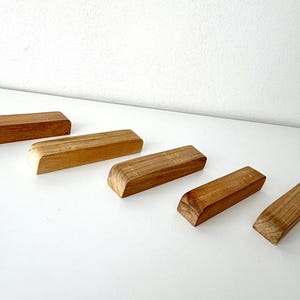 Könnte beinhalten: Fünf rechteckige Holzblöcke mit natürlicher Holzmaserung. Jeder Block hat eine abgeschrägte Kante an einem Ende. Die Blöcke sind diagonal über einer weißen Oberfläche angeordnet und zeigen ihre warmen, erdigen Töne.