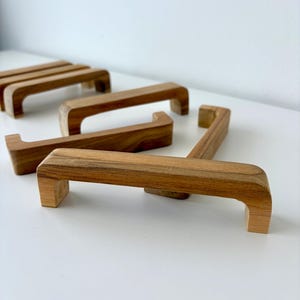 May include: A collection of wooden cabinet pulls, each with a rectangular base and curved handle, are displayed on a white surface. The pulls are made of light brown wood and are approximately 15 cm in length.