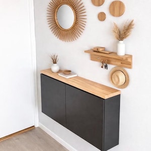 Op de afbeelding: Een moderne entree met een zwarte kast met een houten blad, een zonne-spiegel en houten wanddecoraties. Een klein plankje biedt plaats aan sleutels en een strohoed. Een vaas met gedroogde bloemen voegt een natuurlijk tintje toe.