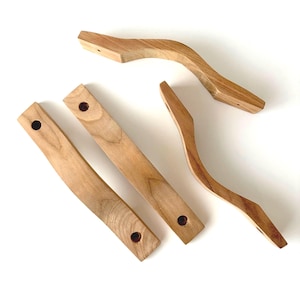 Può includere: Tre maniglie per armadietti in legno marrone chiaro con finitura a venature naturali. Due maniglie sono dritte con bordi arrotondati e due fori preforati, mentre una maniglia presenta una forma curva con due fori di montaggio preforati.