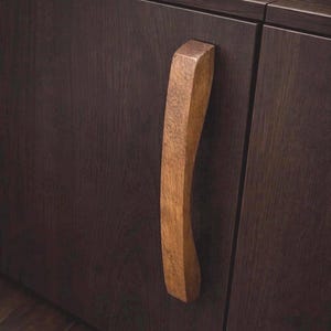 May include: Close-up of a dark brown cabinet door with a light brown, curved wooden handle. The handle is vertically oriented and has a natural wood grain texture. The cabinet door is made of dark brown wood.