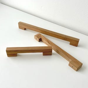 Könnte beinhalten: Drei rechteckige Schrankgriffe aus Holz mit einer natürlichen hellbraunen Holzmaserung. Jeder Griff hat ein gerades Stangendesign mit zwei Montageblöcken an den Enden, wobei sichtbare Schraubenlöcher an den Seiten der Blöcke zu sehen sind.