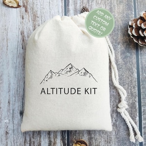May include: A white cotton drawstring bag with a black mountain range illustration and the text "ALTITUDE KIT". A green circle with the text "ADD ANY CUSTOM TEXT OR QUOTE!" is in the upper right corner of the image.