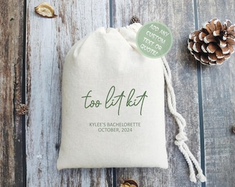May include: A beige drawstring bag with the text "too lit kit" in green script. Below, it reads "Kylee's Bachelorette October, 2024." A green circle with text is on the bag. The bag is on a wooden surface.