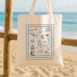 May include: Cream-colored tote bag featuring a blue Cape Cod design with illustrations of a whale, lighthouse, lobster, and other coastal elements. The text "Cape Cod" and "Hannah's Bachelorette" are included in the design. The bag has two cream-colored handles.