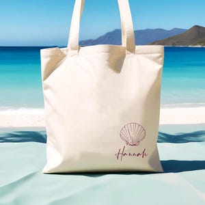May include: A white canvas tote bag with a pink seashell design and the name "Hannah" printed on the front.