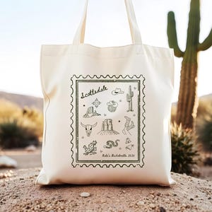 May include: A white canvas tote bag with a black and green design featuring a stamp with the word "Scottsdale" and various illustrations of cacti, cowboy boots, a cowboy hat, a snake, and a skull. The text "Katie's Bachelorette 2025" is printed below the stamp.