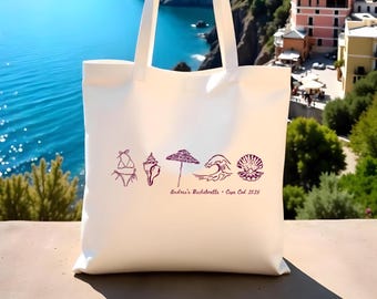 Puede incluir: Un bolso de tela blanco con un estampado gráfico morado de un bikini, una caracola, una sombrilla de playa, una ola y una almeja abierta con una perla. Debajo de los iconos, el texto dice: Andrea's Bachelorette - Cape Cod 2025.