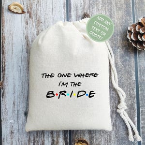 May include: A white drawstring bag with the text "The One Where I'm The B.R.I.D.E." in black with colorful dots. The bag is on a wooden surface.