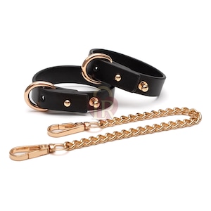 Könnte beinhalten: Zwei schwarze Lederhalsbänder mit goldenen Metallbeschlägen und einer goldenen Kette mit zwei Verschlüssen.