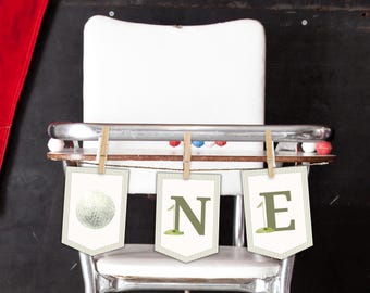 May include: A decorative banner featuring three white pennants with thin vertical stripes. The first pennant displays a golf ball, while the second and third pennants feature the letters N and E in olive green, each accented with a small golf flag.