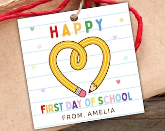 May include: A square gift tag with a white background featuring blue horizontal lines, small pastel hearts, and a yellow pencil shaped into a heart. The text reads HAPPY, FIRST DAY OF SCHOOL, and FROM, AMELIA in colorful letters.