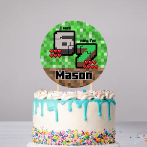 Puede incluir: Un adorno redondo para pastel con el texto "I was 6 now I'm 7" y el nombre "Mason" en un diseño pixelado. El adorno está colocado sobre un pastel blanco con goteo de glaseado azul y chispas coloridas. Es un artículo festivo.