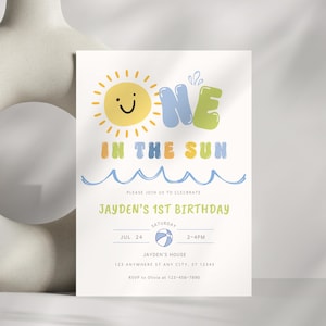 Peut inclure: Une carte d'invitation d'anniversaire rectangulaire avec un soleil jaune souriant, des lettres bulles bleues et vertes formant ONE, et le texte IN THE SUN. En dessous, des vagues bleues, les détails de l'anniversaire de Jayden et un ballon de plage.