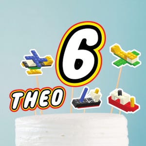 May include: Cake toppers with a construction theme. The toppers include a large number 6, the name "THEO", and miniature toy block airplanes, helicopters, and boats. The toppers are on wooden sticks and are placed on top of a white cake.