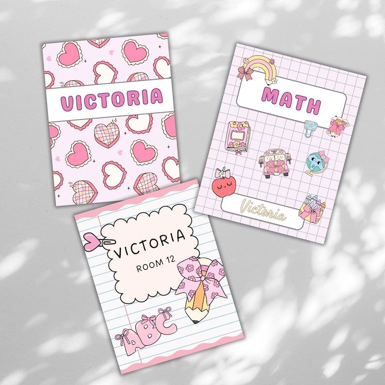 May include: Three personalized notebooks with pink covers. One features a heart pattern and the name "Victoria." Another says "Math" with cute illustrations. The third notebook has "Victoria, Room 12" and "ABC" text.
