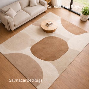 Op de afbeelding: Een beige en bruin vloerkleed met een abstract ontwerp met grote, afgeronde vormen. Het kleed ligt op een houten vloer, met een beige bank, een ronde houten salontafel en een plant in een pot op de achtergrond.