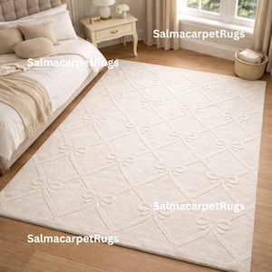 Può includere: Tappeto rettangolare color crema con un motivo a fiocco ripetuto. Il tappeto è posato su un pavimento in legno in una camera da letto. Il design presenta fiocchi in rilievo e strutturati all'interno di una griglia.