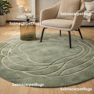 May include: A round, sage green rug with a cream-colored, wave-like pattern. The rug is placed on a wooden floor, with a beige armchair and a glass-topped table with a brown base in the background. The rug's diameter is approximately 7 feet.