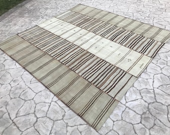 May include: A large, rectangular rug with a patchwork design. The rug features alternating rows of light beige and off-white panels, each with vertical stripes in varying shades of brown and black. The rug is made of a woven material.
