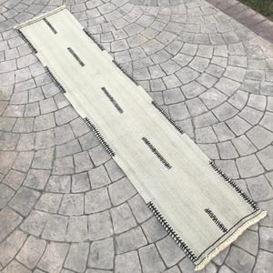 May include: A long, off-white runner rug with black stitching and fringe. The rug has a simple geometric pattern.