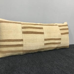 May include: Rectangular beige pillow with brown horizontal stripes. The pillow has a textured surface and is placed on a dark gray surface. The pillow is a decorative item.