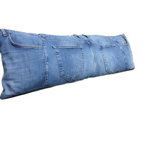May include: A long, rectangular cushion made from repurposed denim jeans. The cushion features the pockets and seams of the jeans, with a light blue colour. The cushion is designed for decorative or functional use.
