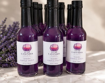 Może przedstawiać: Kilka przezroczystych szklanych butelek wypełnionych fioletowym syropem lawendowym. Każda butelka posiada białą etykietę z napisem Lush Lavender Farm, lavender simple syrup oraz 5 fl. oz. / 150 ml. Butelki mają czarne nakrętki.