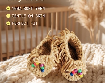 May include: A pair of tan crocheted baby booties featuring fringe detailing and colourful plastic beads on the front. The text on the image reads: Handcrafted comfort for tiny toes, 100% SOFT YARN, GENTLE ON SKIN, PERFECT FIT, TINY TOES, Handmade, Cozy.