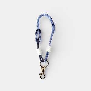 May include: A wrist lanyard featuring a blue and white patterned rope loop, a solid dark blue rope section with a knot, two white rope accents, and a gold-toned metal lobster clasp attached to a circular metal ring at the base of the cord.