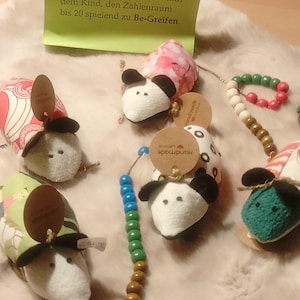 Könnte beinhalten: Fünf Stoffmäuse mit weißen Gesichtern und schwarzen Ohren, jede mit einem gemusterten Rücken. Zwei Mäuse haben Schnüre mit bunten Holzzählperlen. Ein grünes Schild mit dem deutschen Text Rechenmäuse beschreibt den Zahlenraum bis 20.