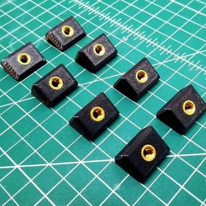 May include: Nine black plastic corner brackets with gold-colored threaded inserts. The brackets are arranged in a staggered pattern on a green cutting mat.