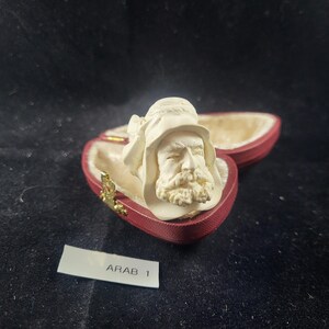 May include: A cream-colored bust of a man with a beard and hat, presented in a heart-shaped red box with a gold clasp. The box is open, revealing a soft, beige interior. The label reads "ARAB 1".